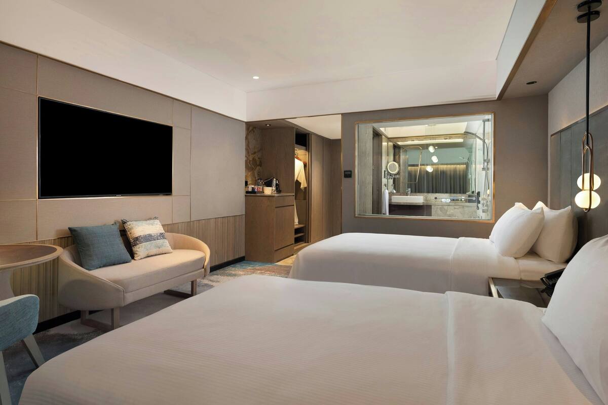 A modern hotel room with two beds, a flat-screen TV, a seating area with a table, and a bathroom visible through a large mirror.