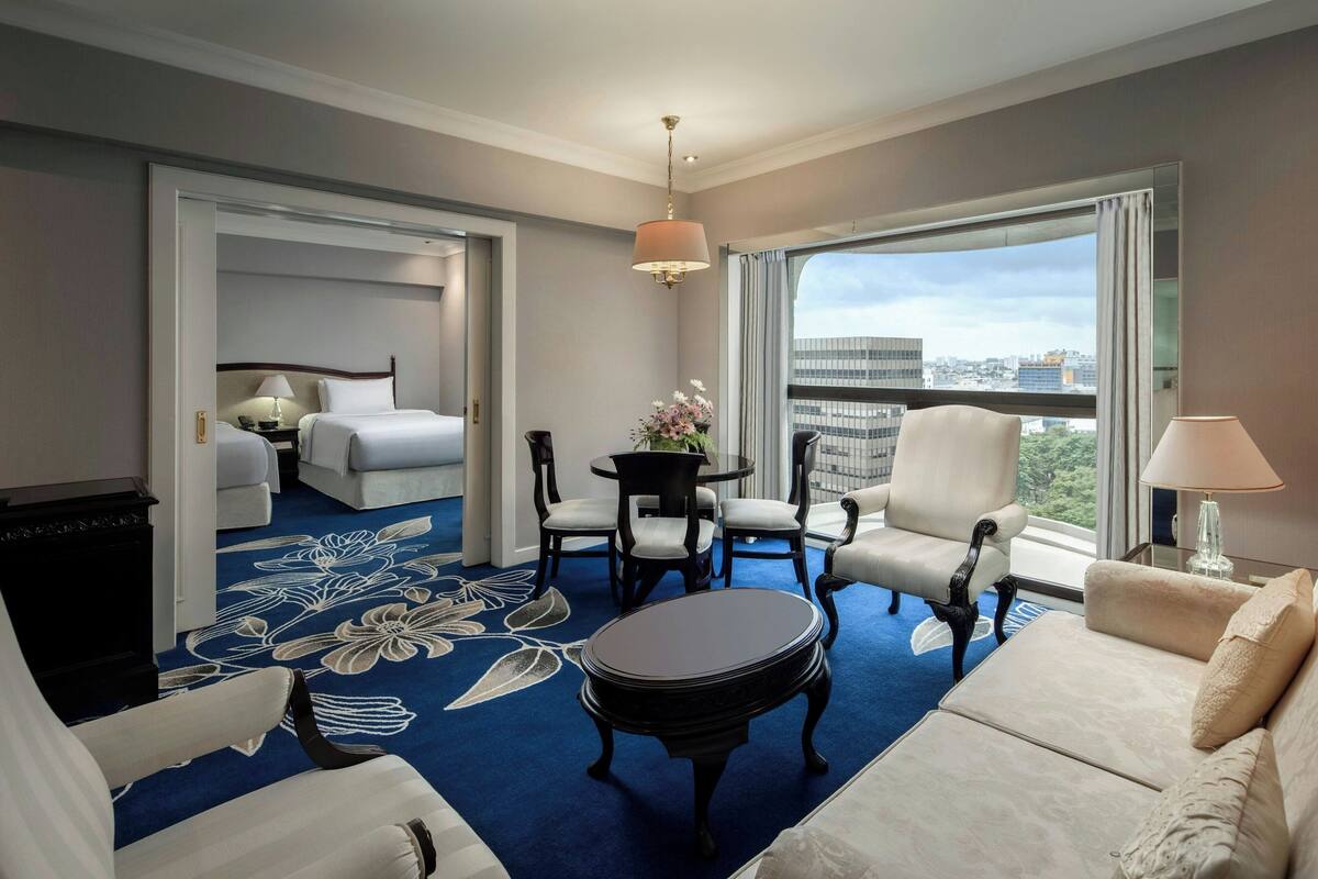 A hotel room with a bed, a sofa, a dining table with chairs, a small round table with a lamp, and a large window with a city view.