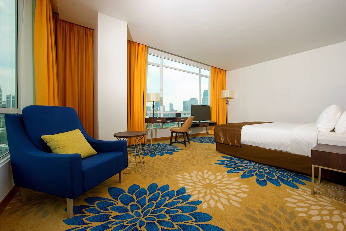 Een hotelkamer met een groot raam, een blauwe fauteuil, een bed, een bureau en uitzicht op de stad.