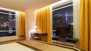 Habitación de hotel con una cama grande, un escritorio con ordenador, una silla y televisor. La habitación tiene vistas a un horizonte urbano nocturno.