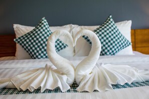 Una cama impecablemente hecha con una toalla dispuesta en forma de corazón y almohadones a cuadros.