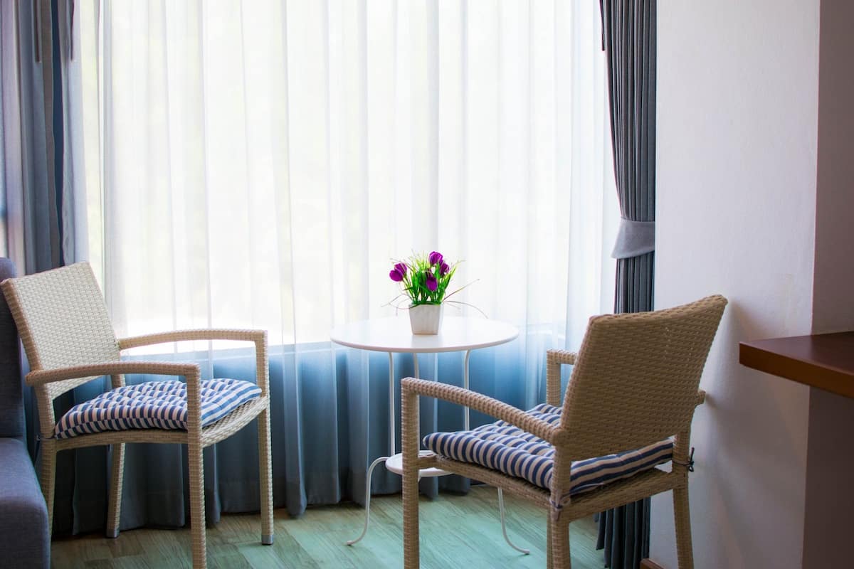 A room with two wicker chairs, a small round table with a potted plant, and a view through sheer curtains.