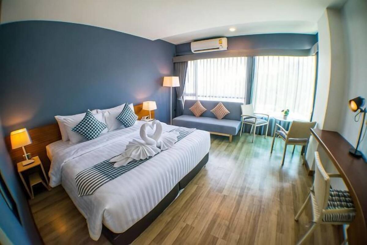 A hotel room with a bed, bedside lamps, a desk, a chair, a sofa, and a window with blinds.