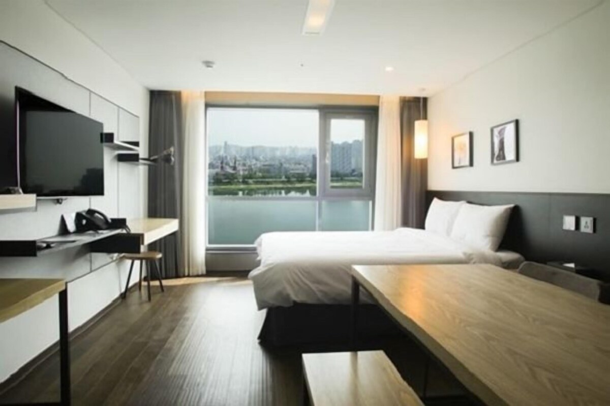 A modern hotel room with a bed, a desk, a flat-screen TV, and a large window offering a city view.