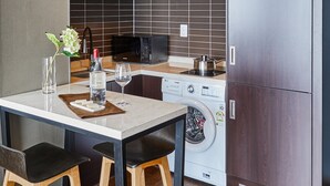 A compact kitchen with a microwave, washing machine, and refrigerator. There is a small dining table with two chairs and a vase with flowers. - Hotel Riverside (Ulsan)