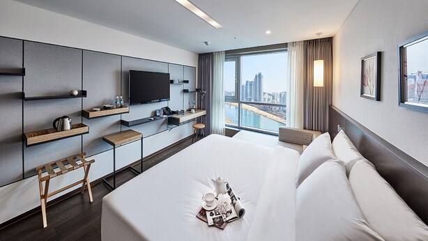 A modern hotel room with a large bed, a flat-screen TV, a desk with a chair, and a city view. - Hotel Riverside (Ulsan)