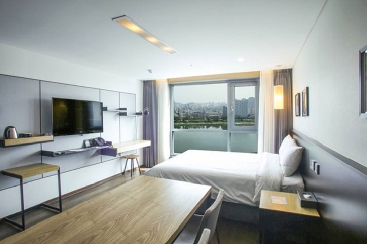 Una habitación de hotel moderna con una cama grande, un escritorio con televisor de pantalla plana y vistas de la ciudad a través de amplias ventanas, con un paisaje urbano visible desde el exterior.