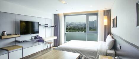 A modern hotel room with a large bed, a desk with a flat-screen TV, a view of the city through large windows, and a cityscape visible outside.