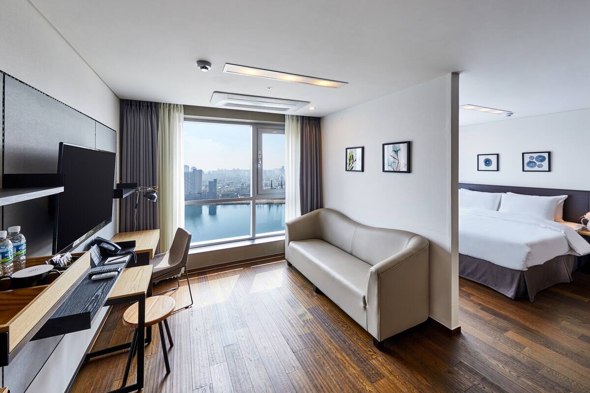 Habitación de hotel con escritorio, silla, sofá, cama y grandes ventanales con vistas a la ciudad.