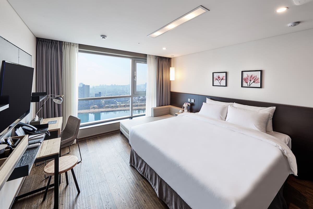 Habitación de hotel con una cama grande, un escritorio con computadora, una silla y vista a un paisaje urbano a través de un ventanal amplio.