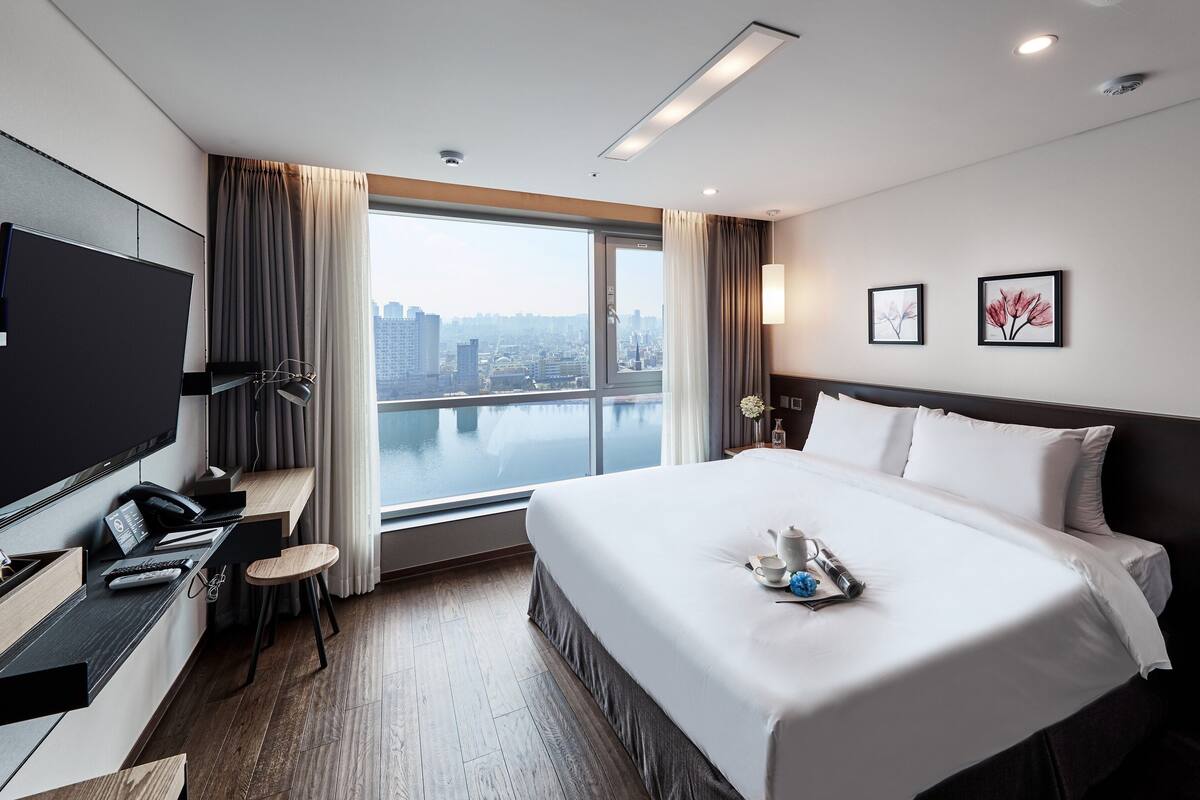 Una habitación de hotel moderna con una cama grande, un televisor de pantalla plana, un escritorio con silla y vistas a la ciudad a través de amplias ventanas.