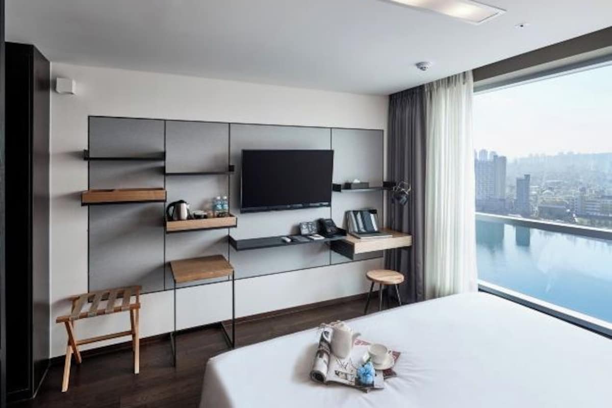 Habitació d'hotel moderna amb un llit gran, un televisor de pantalla plana fixat a la paret, un petit tamboret de fusta i vista a un paisatge urbà a través d'una finestra gran.