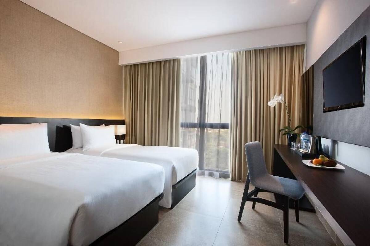 A hotel room with two beds, a desk, a chair, a TV, and a large window with curtains.