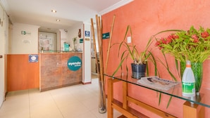 A hotel reception area with a wooden reception desk, a glass table with a newspaper, a bottle of alcohol, and potted plants. - Hotel Calle 112 (Bogotá)