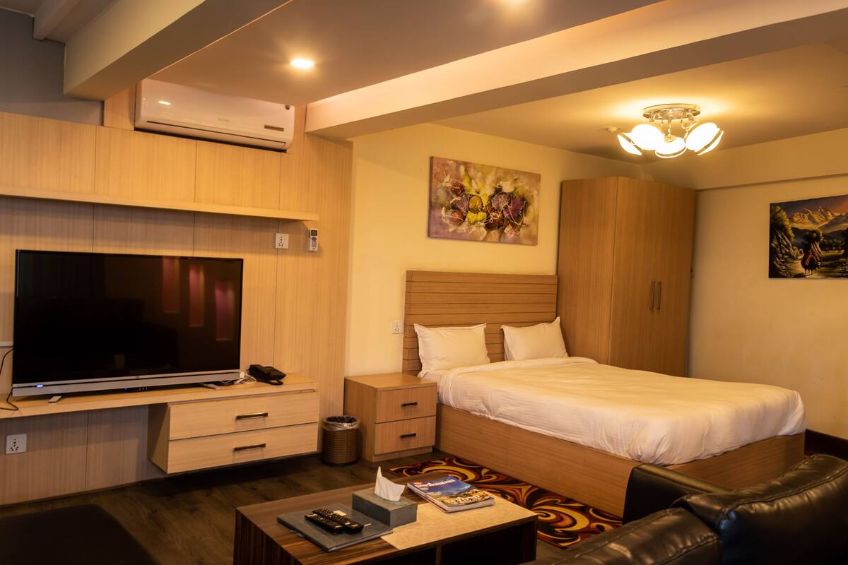 Habitación de hotel con un televisor de pantalla plana grande, cama con ropa blanca, armario de madera, mesita de noche pequeña y alfombra colorida.