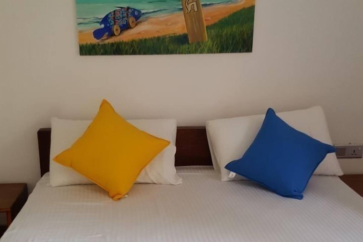 A bed with white bedding, two pillows (one yellow, one blue), and a folded teal towel. A colorful painting of a beach scene hangs above the bed.