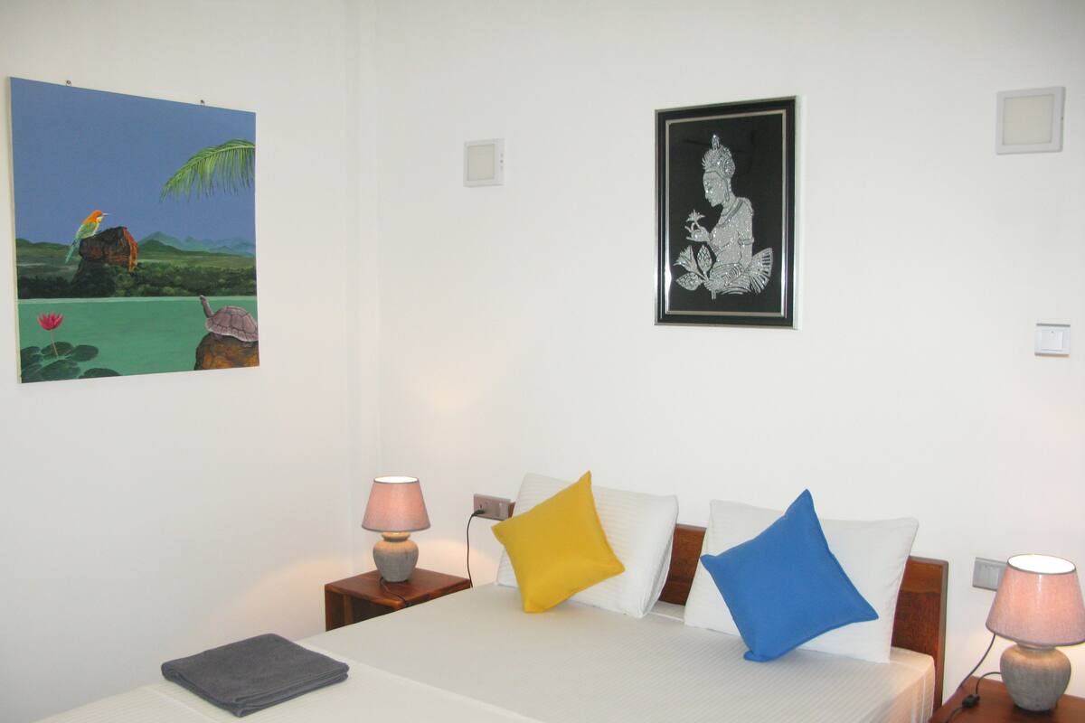 A hotel room with a bed, two bedside tables, a lamp, a painting, and a framed picture on the wall.