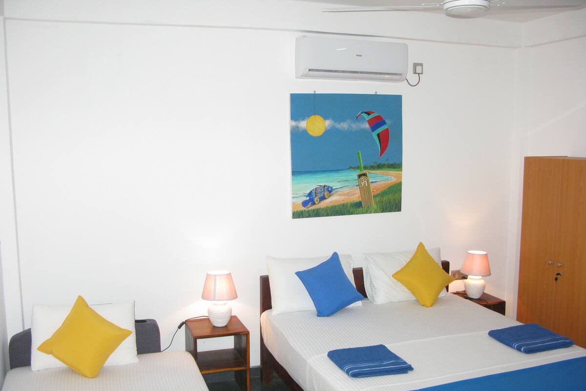 A hotel room with two beds, a painting of a beach scene, an air conditioning unit, and a wooden wardrobe.