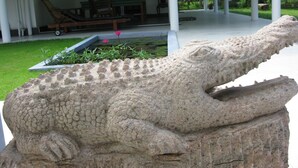 A crocodile sculpture in a garden with a covered seating area and a pond in the background.