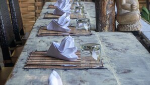 A long table with neatly folded napkins and clear glasses, set against a rustic backdrop with wooden elements and a statue. - Swar Bali Lodge (Munduk)