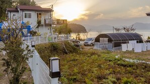 Ein Küstenwohngebiet mit Häusern, einem eingezäunten Garten und Blick auf das Meer bei Sonnenuntergang.