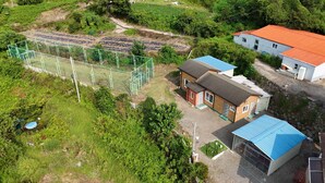 A residential area with a fenced sports field, a house with a blue roof, and a small shed. - Sea and Sunset Pension (Namhae)