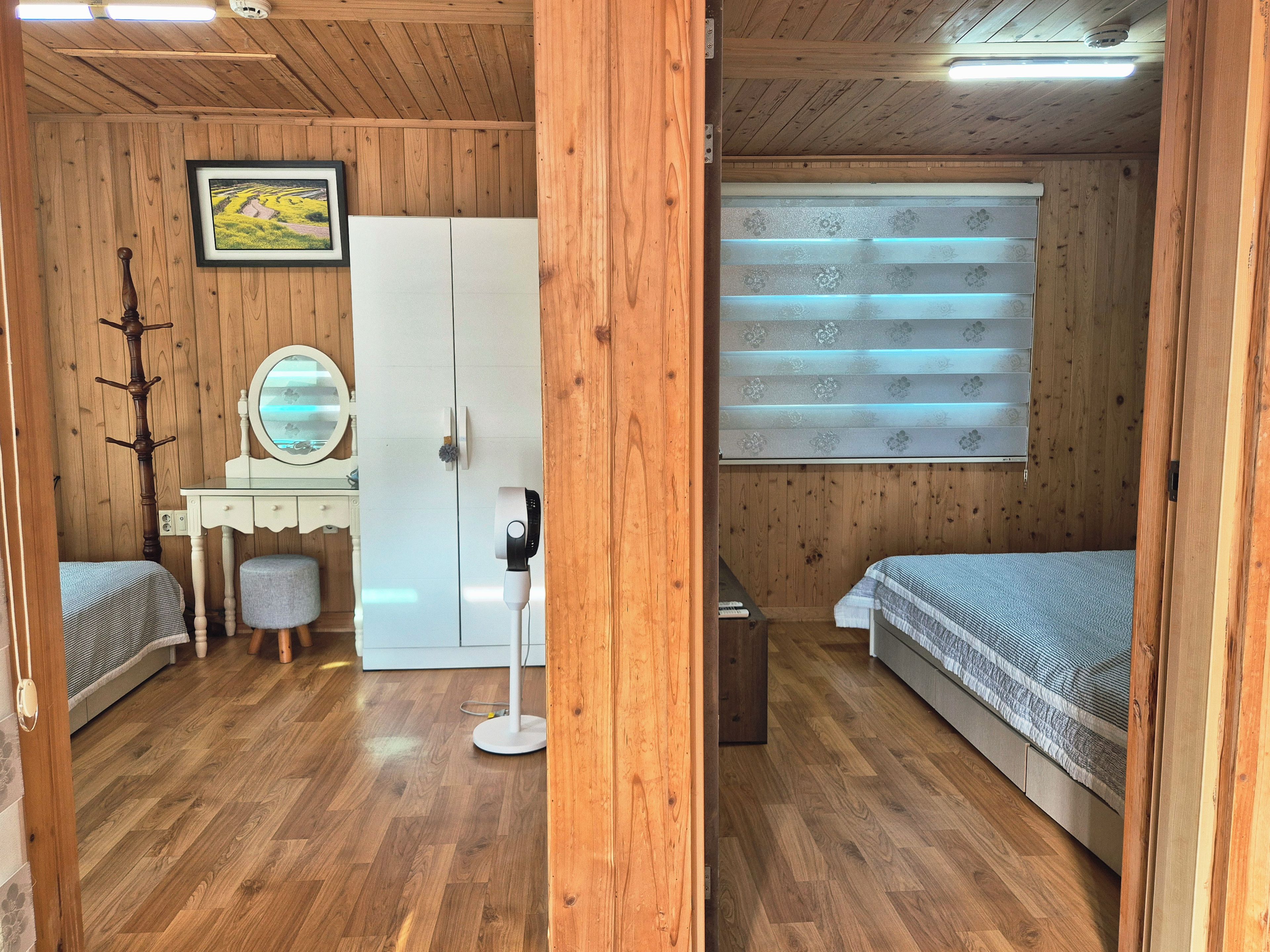 Une pièce aux murs lambrés, comprenant un lit, un bureau avec un miroir, une armoire et une fenêtre avec des stores.