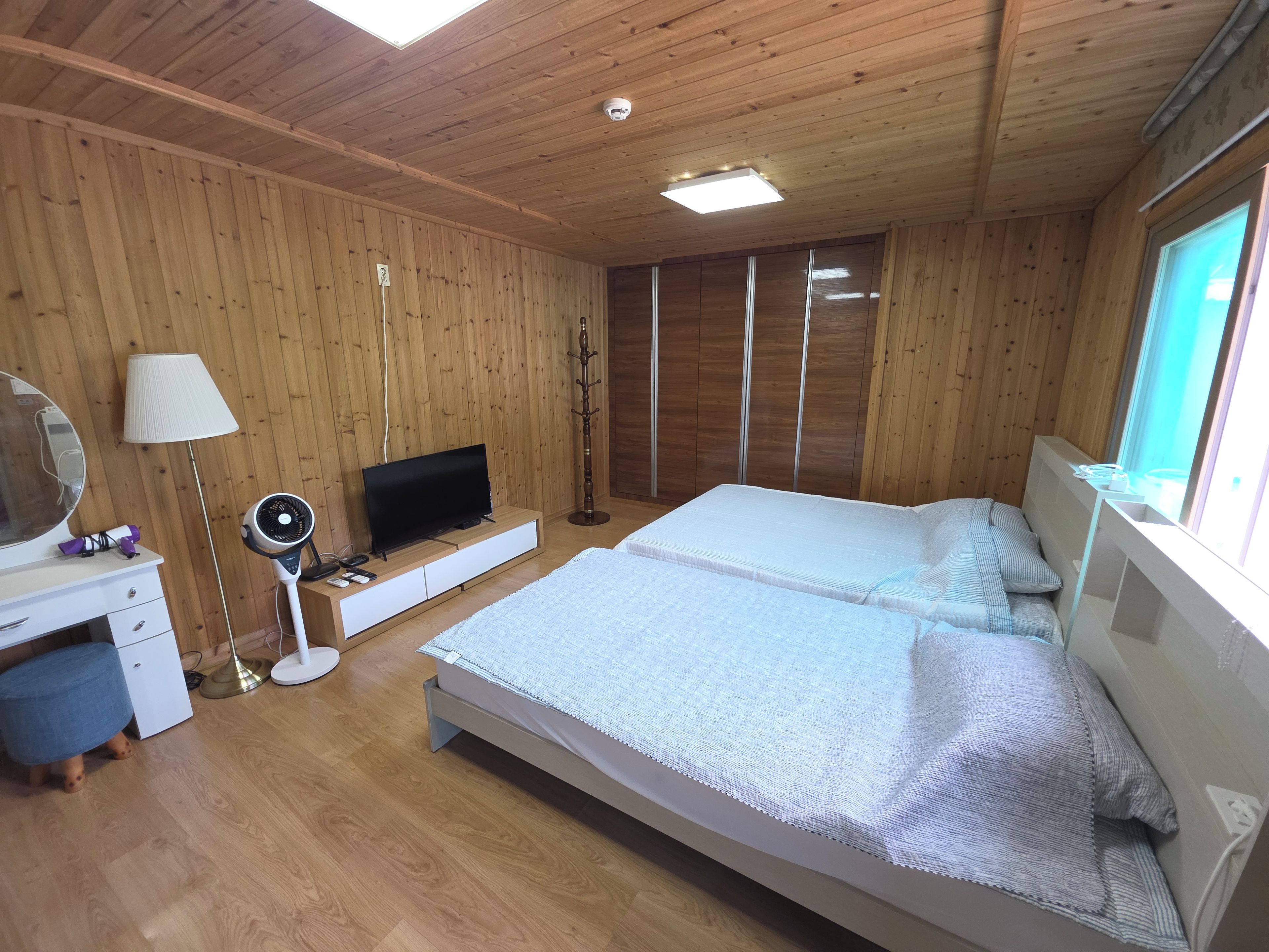 Una habitaci ón con paredes de madera, una cama con colchón y almohadas, un televisor sobre un soporte, una lámpara y un ventilador de pie.