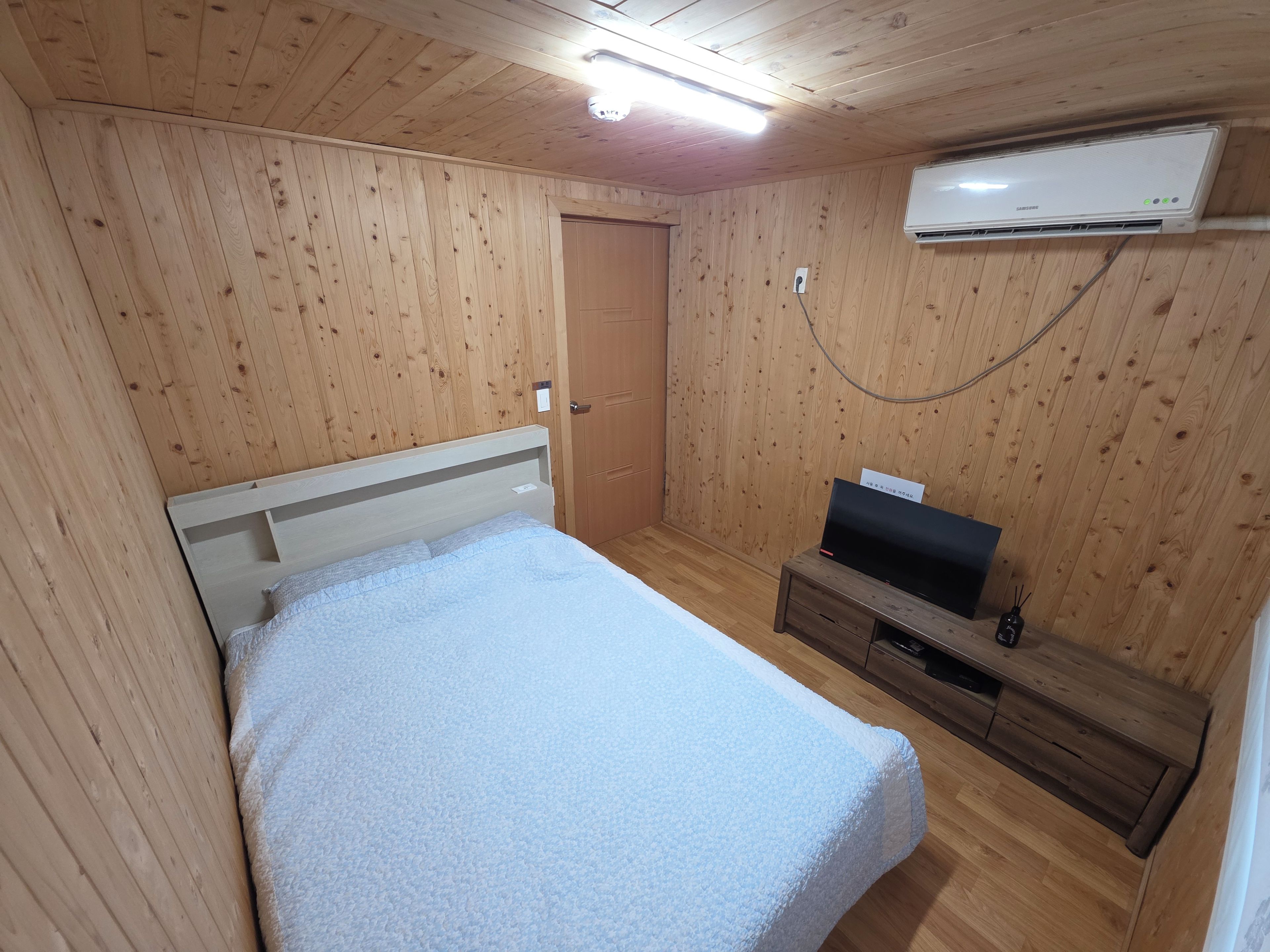 A room with a bed, a TV on a wooden stand, an air conditioner on the wall, and a door.