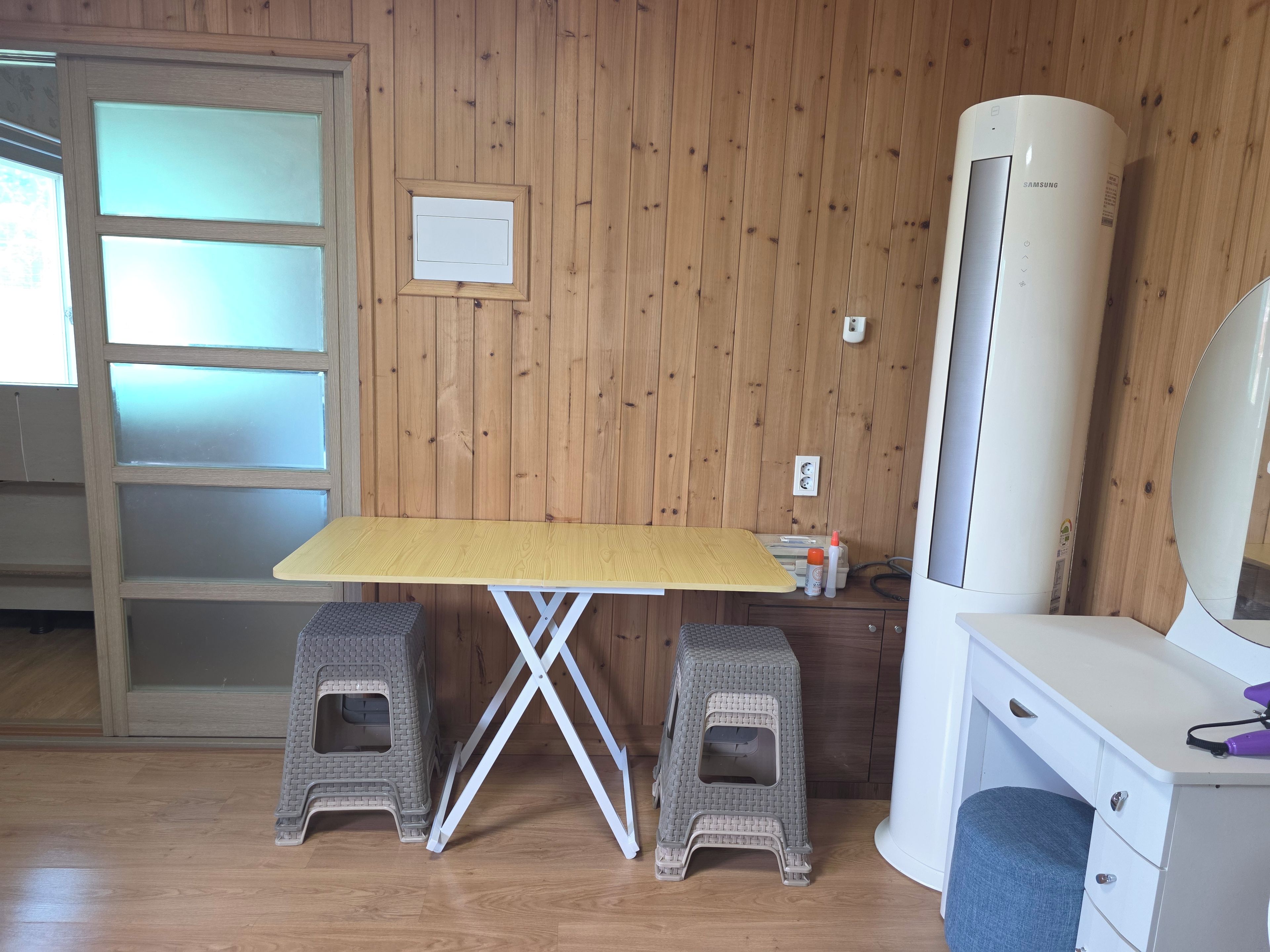 Una habitación con un revestimiento de madera, un escritorio blanco con un taburete, un aparador blanco y un equipo de aire acondicionado grande.