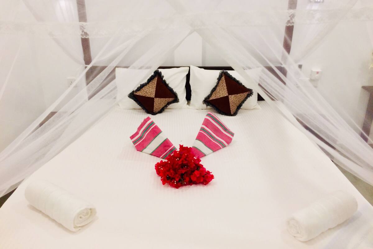 Una cama bien hecha con sábanas blancas, dos almohadones decorativos y un ramo de flores rojas.