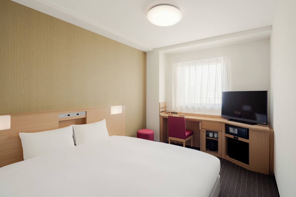 Una habitación de hotel con una cama grande, un escritorio con televisor y una ventana con cortinas traslúcidas.