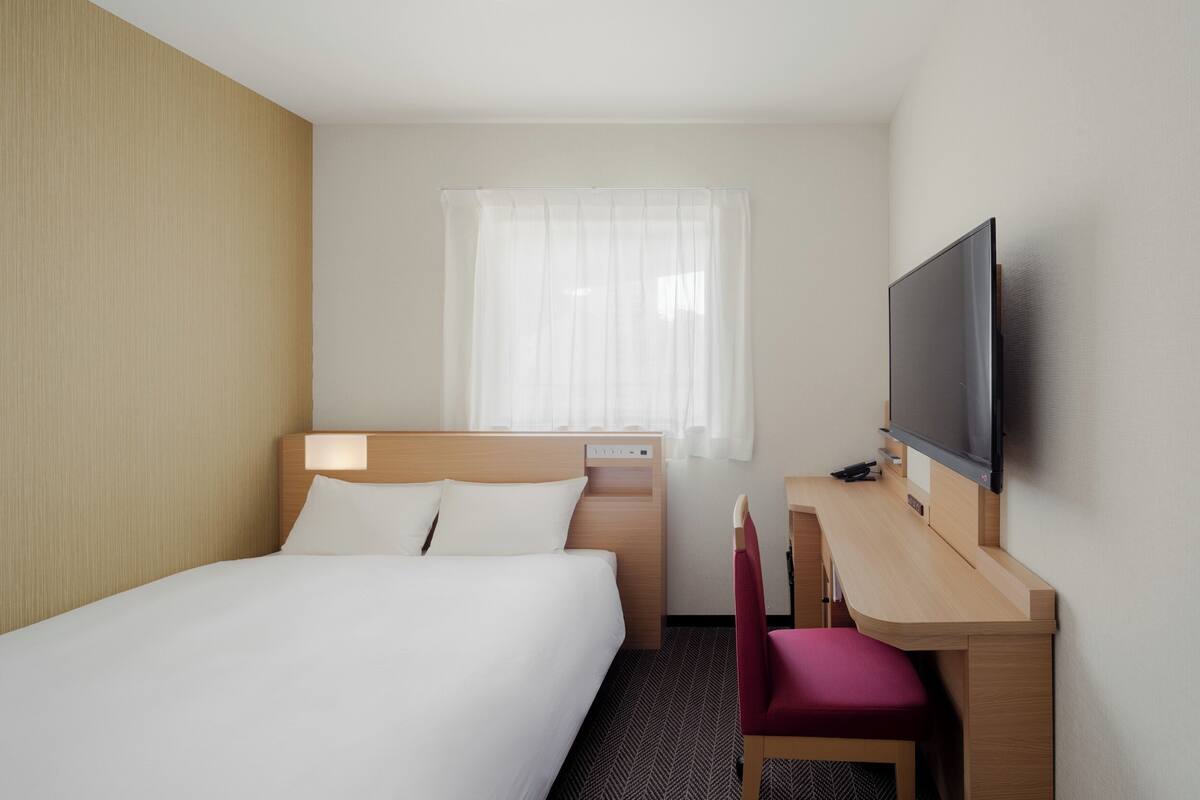 Habitación de hotel con cama, escritorio, silla, televisor y ventana con cortina translúcida.