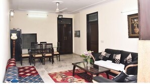 A living room with a sofa, coffee table, and dining area with chairs and a table. There is a ceiling fan and a framed picture on the wall. - Jagannath Guest House (New Delhi)