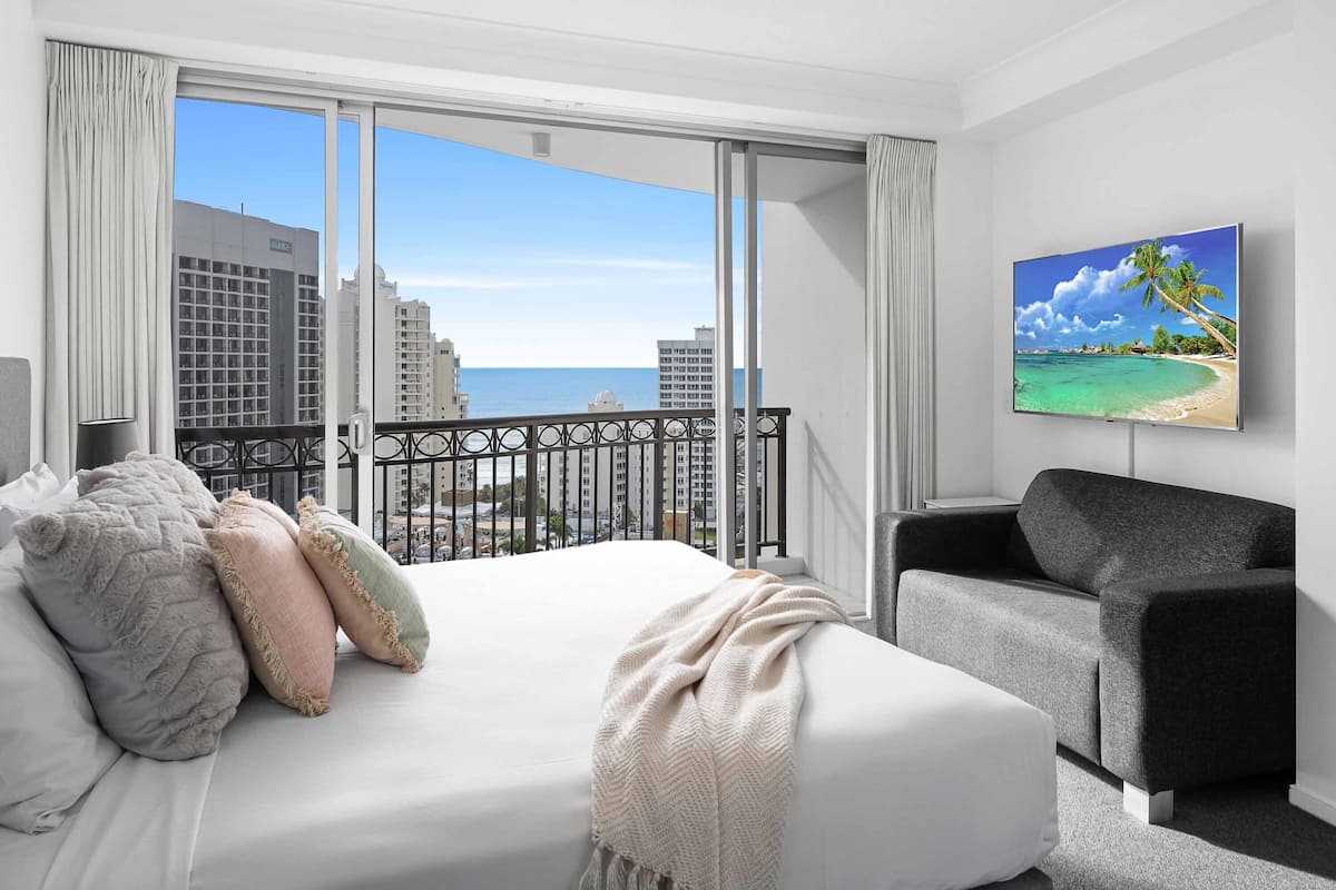 A hotel room with a bed, a grey armchair, a large window with a view of the ocean and buildings, and a painting of a beach scene.