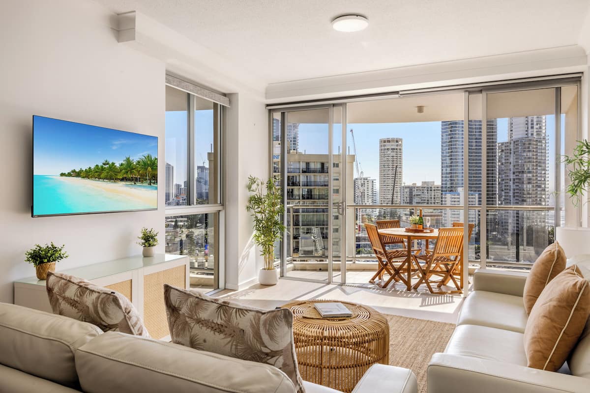 A modern living room with a sofa, a coffee table, and a dining area with chairs and a table. There is a large window with a city view and a painting of a beach on the wall.