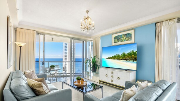 A modern living room with a large TV, comfortable seating, a balcony with a view of the ocean, and a chandelier. - Holiday Holiday Chevron Renaissance (Surfers Paradise)