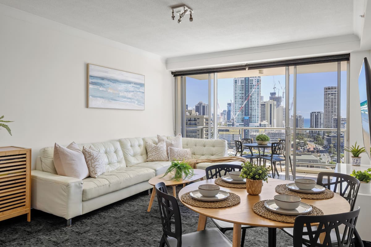 A modern living room with a white sofa, a dining table with four chairs, and a large window overlooking a cityscape.