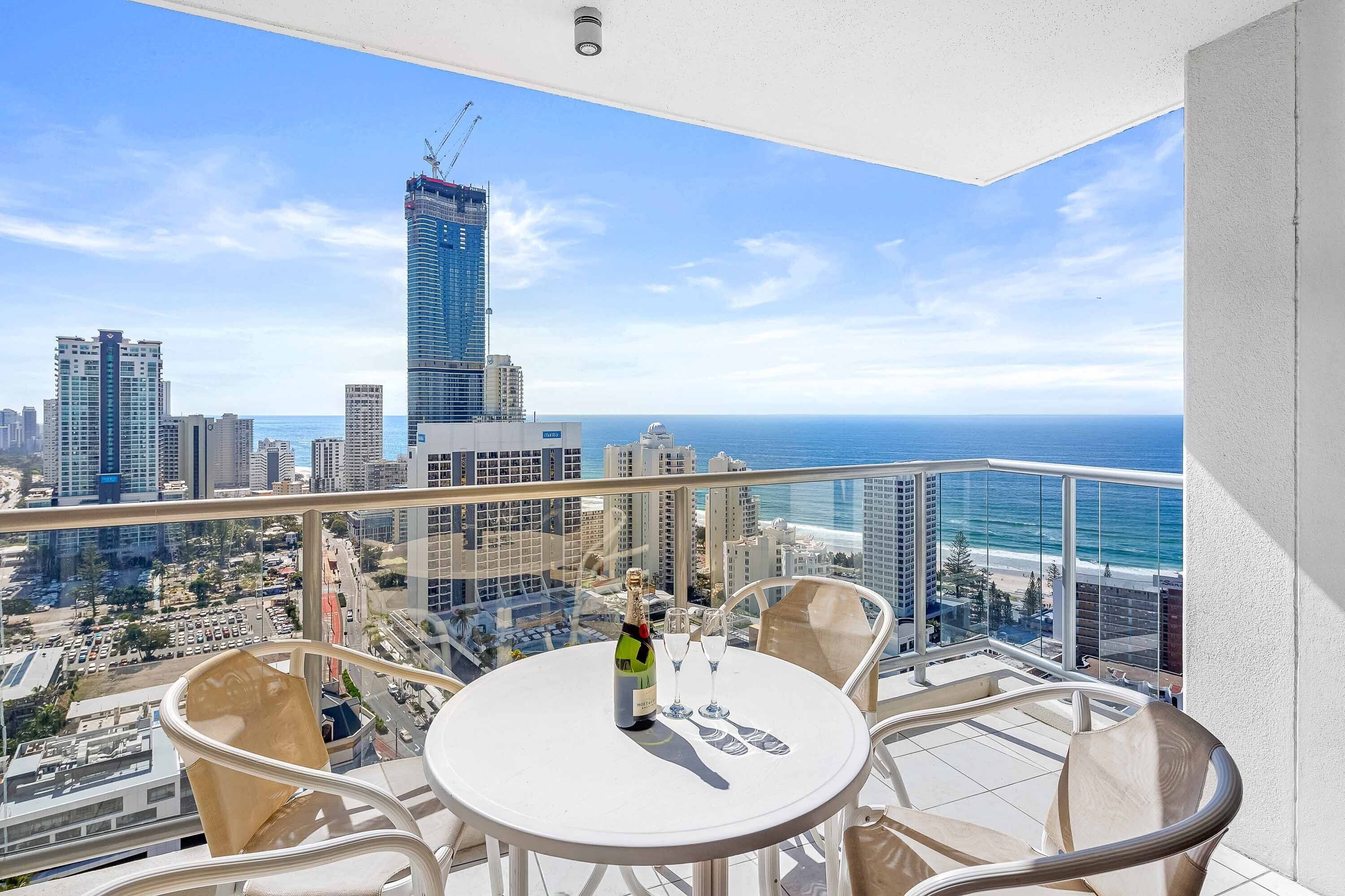 A balcony with a table, chairs, and a bottle of champagne. The balcony overlooks a cityscape with tall buildings and a view of the ocean.