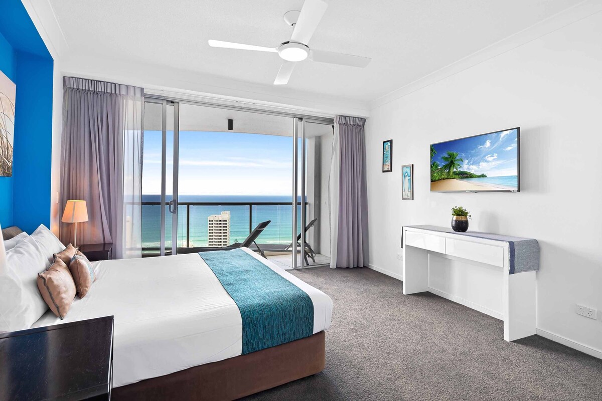 A hotel room with a bed, a desk, a chair, a TV, and a view of the ocean.