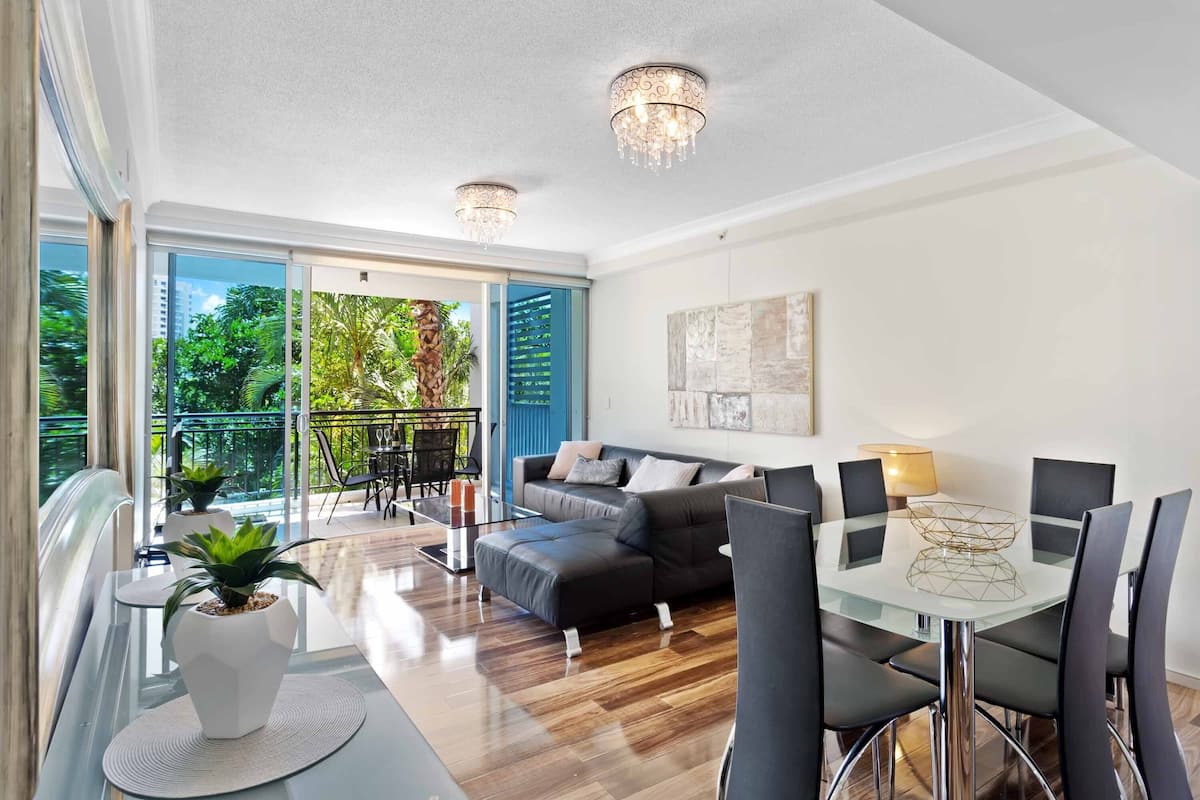 A modern living room with a glass dining table, black leather sofa, and a balcony with a view of greenery.