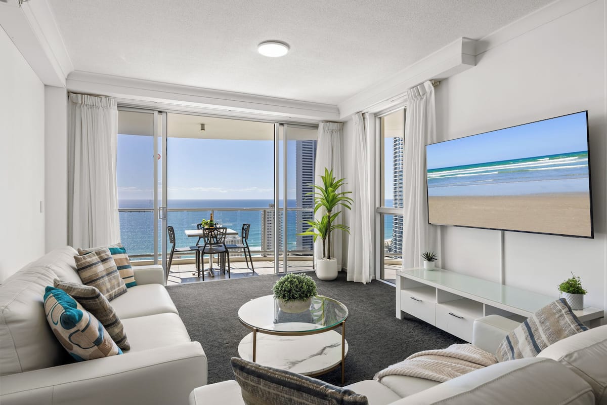 A modern living room with a sofa, armchair, and coffee table, a TV on the wall, and a balcony with a view of the ocean.