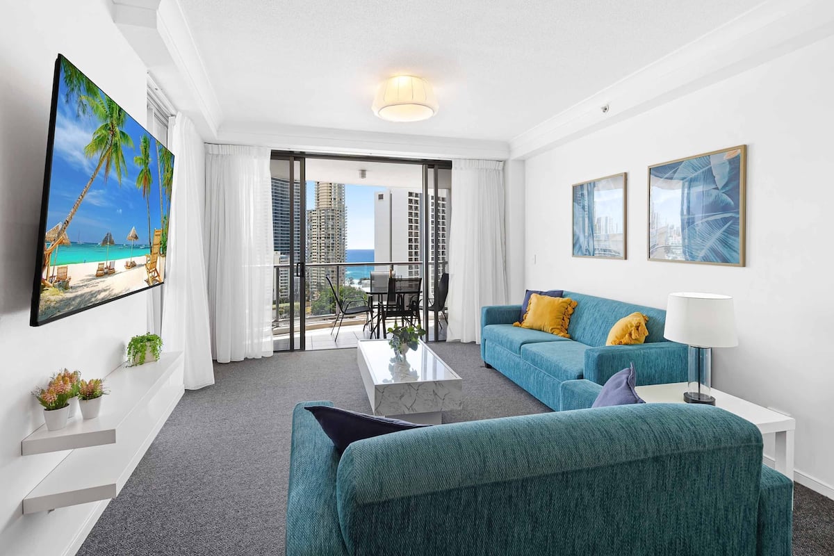 A modern hotel room with a large balcony, a sofa, a coffee table, and a TV mounted on the wall.