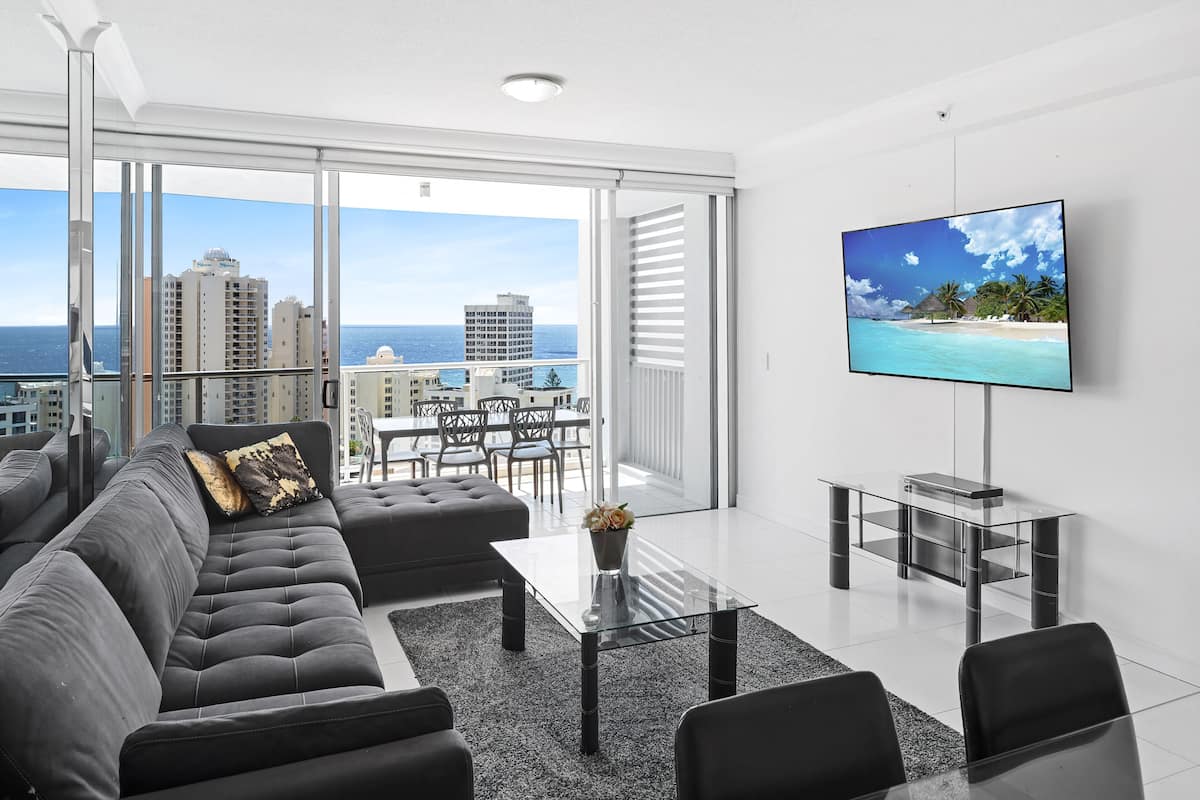 A modern living room with a large TV, a sofa, a coffee table, and a view of the ocean and buildings.