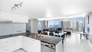 A modern kitchen with a dishwasher, a dining area with a table and chairs, and a view of the city through large windows. - Holiday Holiday Chevron Renaissance (Surfers Paradise)