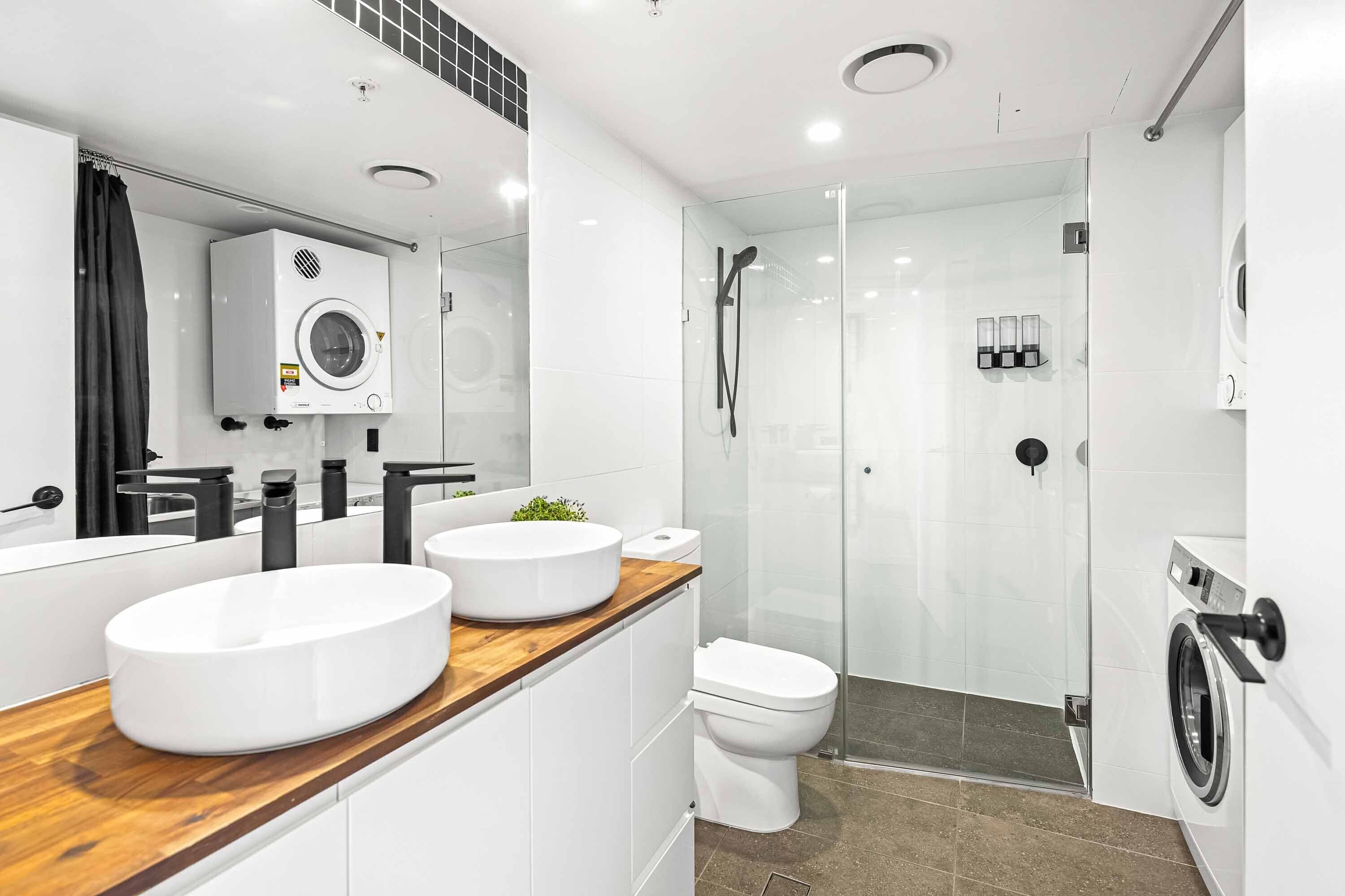 Un bany modern amb un lavabo de fusta, una rentadora integrada, un espai de dutxa amb envàs de vidre i un vàter.