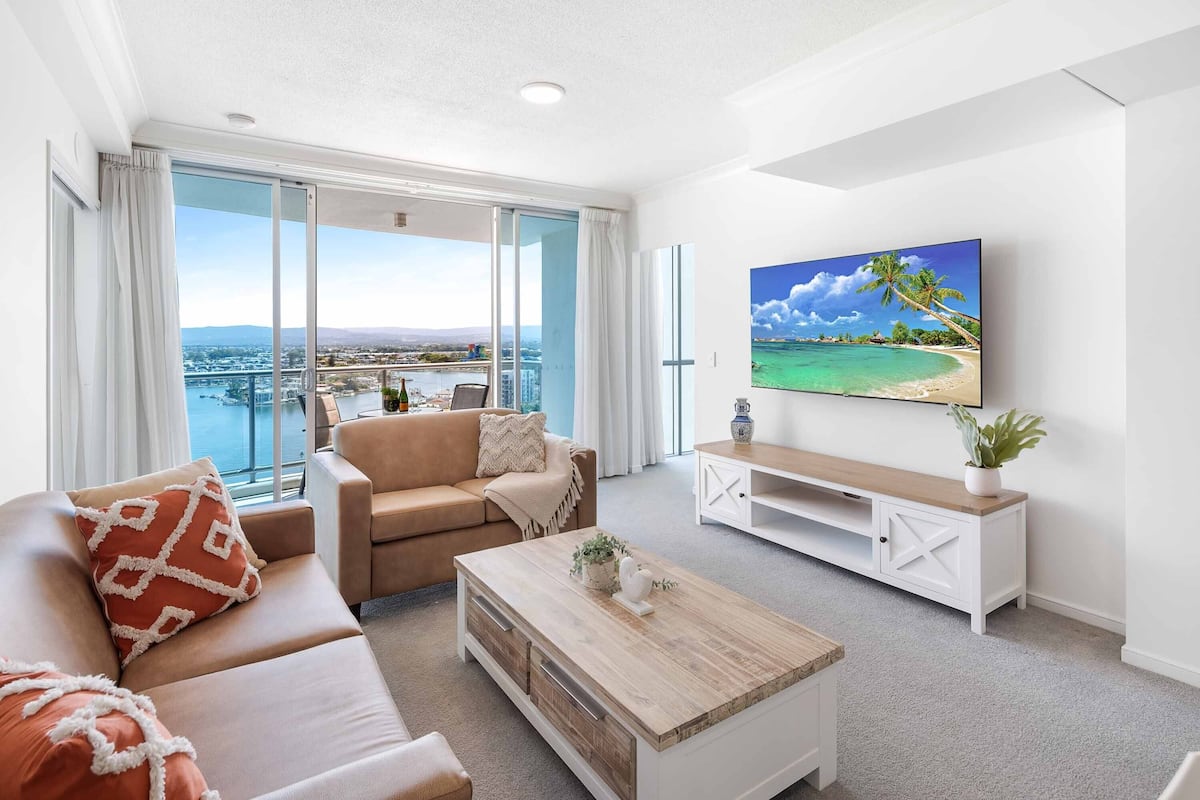A modern living room with a large sliding glass door leading to a balcony, a beige sofa, a wooden coffee table, and a flat-screen TV mounted on the wall.