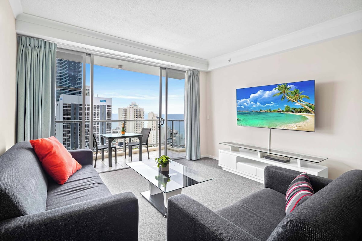 A modern living room with a large TV, grey sofas, a glass coffee table, and a balcony with a city view.