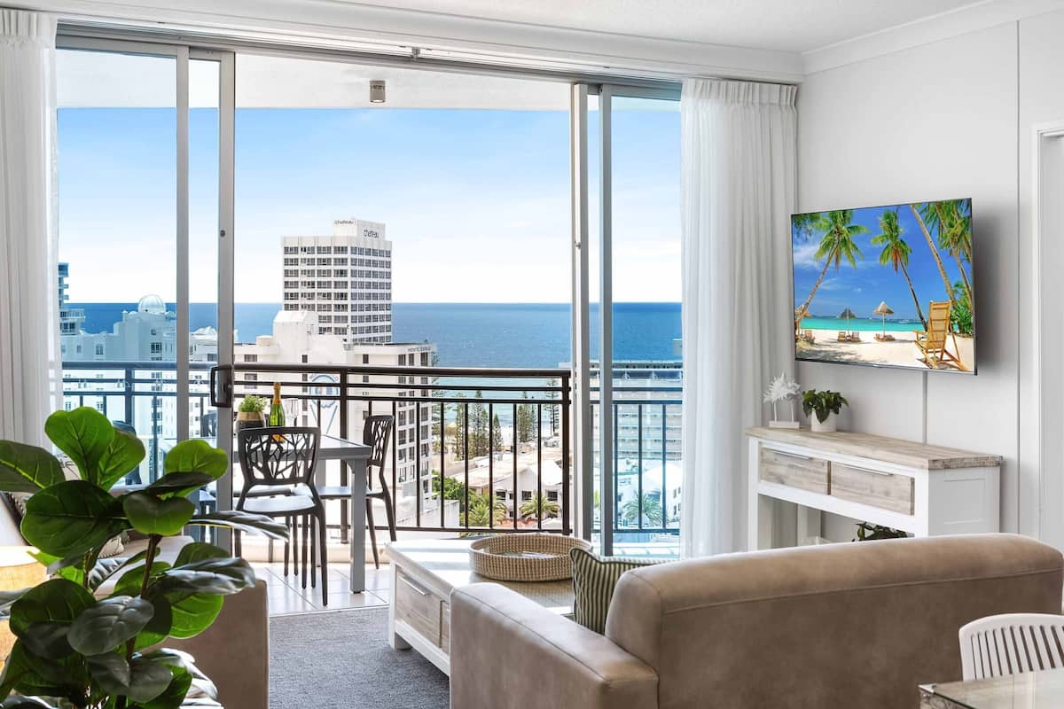 A modern hotel room with a balcony, large windows, and a view of the sea and buildings.
