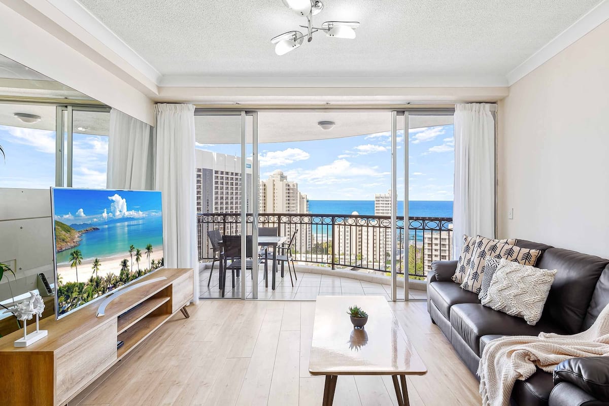 A modern living room with a large TV, a sofa, a coffee table, and a balcony with a view of the sea and buildings.